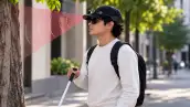 Ilustración generada con inteligencia artificial de un joven con lentes oscuros que utiliza un bastón blanco y una gorra negra equipada con sensores. Frente a él aparece un haz rojizo que representa la detección de obstáculos.