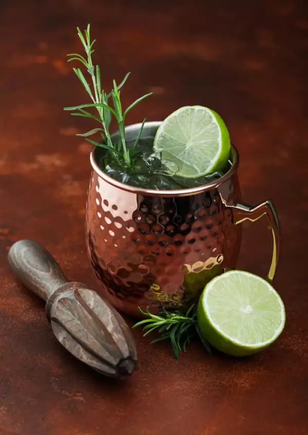 Moscow mule en una taza de cobre con lima y romero y exprimidor de madera sobre fondo de mesa de cocina marrón