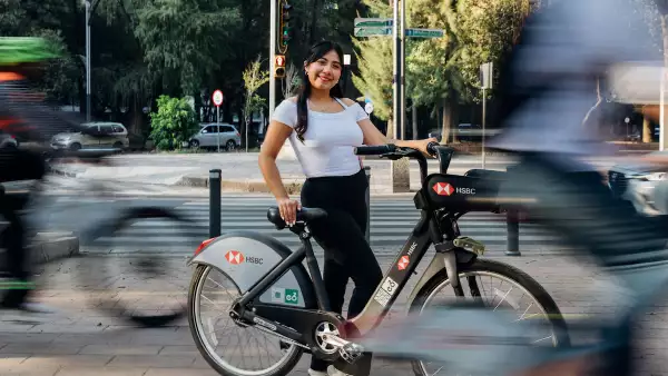 Una joven tomando una Ecobici posa para la foto mientras en el fondo se ve la silueta barrida de otros ciclistas que pasan.    