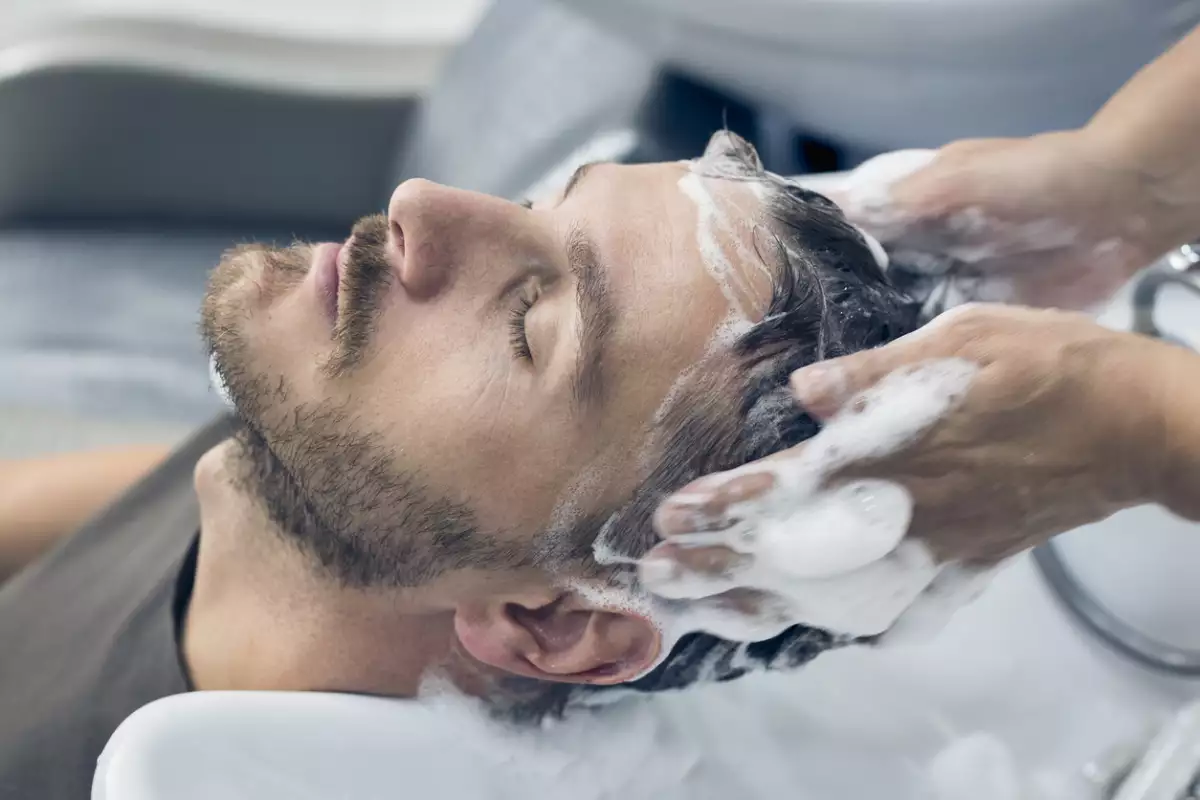 Foto de un hombre joven con los ojos cerrados mientras le lavan el cabello en una peluquería con espuma abundante.