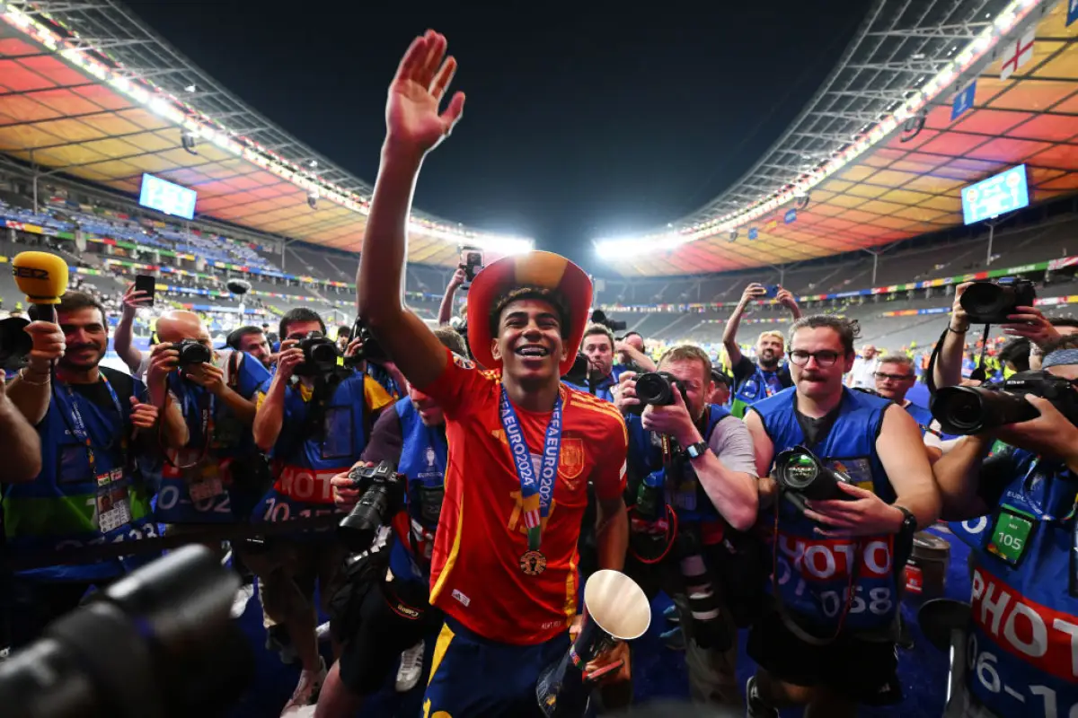 Foto de Lamine Yamal al final de la Eurocopa, sonriendo con la camiseta de la selección de España, una medalla de campeón colgando del cuello y la mano levantada en señal de victoria, rodeado de fotógrafos de todo el mundo.