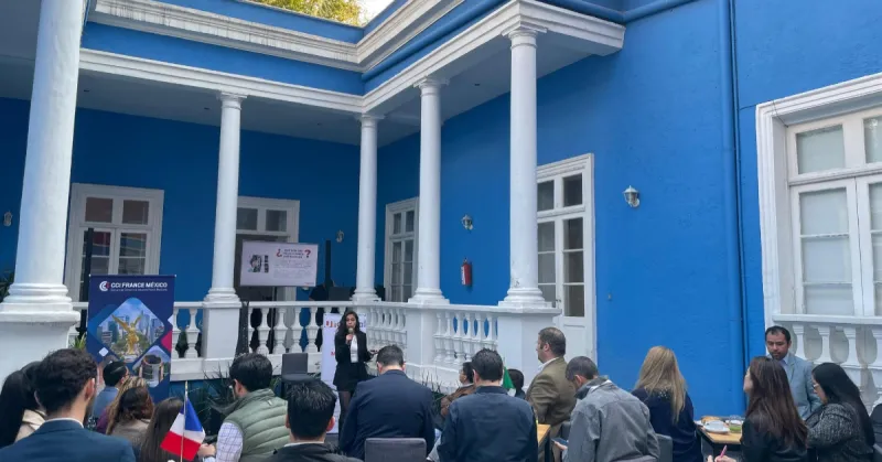 Ventanales y columnas de una casa azul con blanco. Personas sentadas en el exterior frente a una mujer con micrófono.jpg