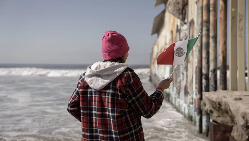 Una persona sostiene una bandera de México con su mano derecha en el muro fronterizo entre México y Estados Unidos.