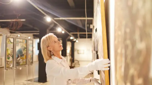 Una galerista coloca una pintura en la pared al interior de una sala de arte.