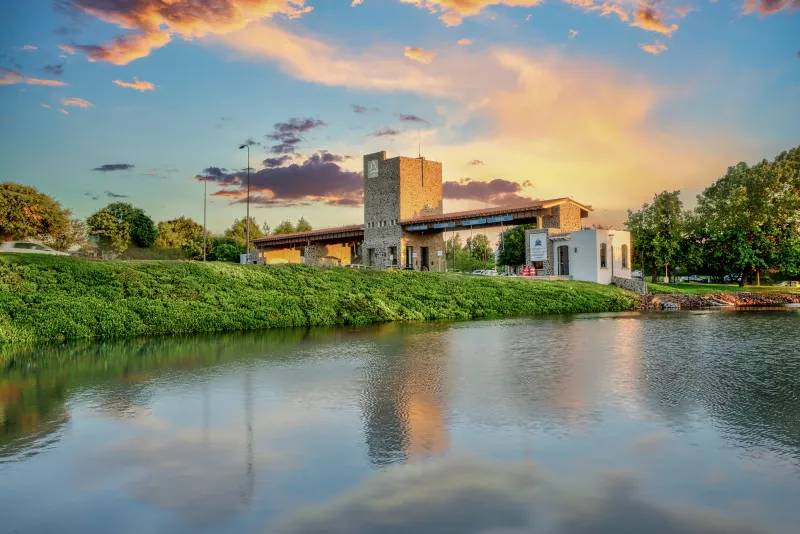 Atardecer en El Campanario Residencial, se observa la Casa Club entre la vegetación a un lado del lago y, de fondo, el cielo con nubes en tono naranja.