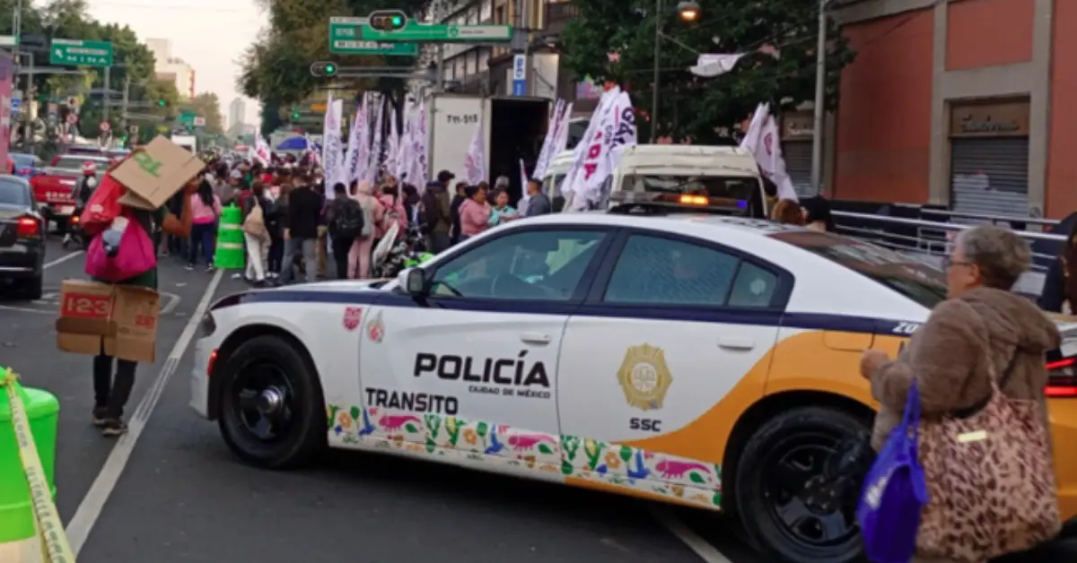 Una patrulla color blanco con vivos amarillos con letras que dice policía y tránsito en una calle con personas y pancartas.jpg