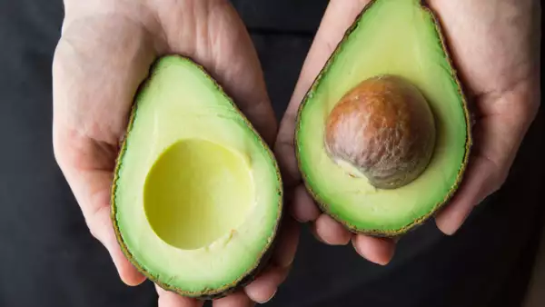 Young Caucasian Woman in Black Apron Holding in Hands Ripe Beautiful Halved Organic Australian Avocado with Pit. Healthy Lifestyle Vegan Vitamin E Oil. Creative Minimalist Food Poster Banner. Kinfolk
