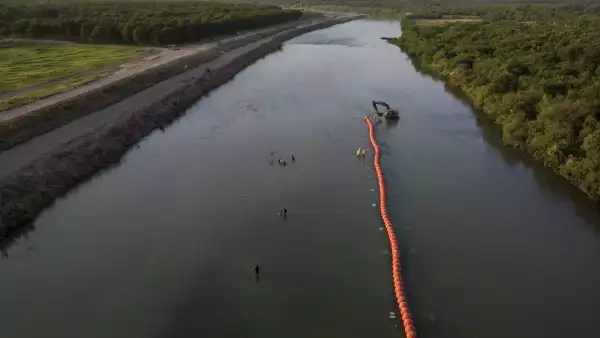 El huerto de nueces de Heavenly Farms se ve a la izquierda mientras las familias migrantes navegan por el río Río Grande pasando por trabajadores y boyas mientras buscan un punto de entrada a los Estados Unidos desde México en Eagle Pass, Texas, EE. UU., el 27 de julio de 2023.
