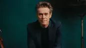 Foto del actor Willem Dafoe con playera negra, sin barba y saco verde oscuro