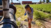 Niña se moja con agua que sale de una tubería en un campo agrícola, mientras un tren de carga circula al fondo.
