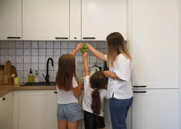 Rear view of a mother with two daughters, in the kitchen at home, daughters reaching for grapes