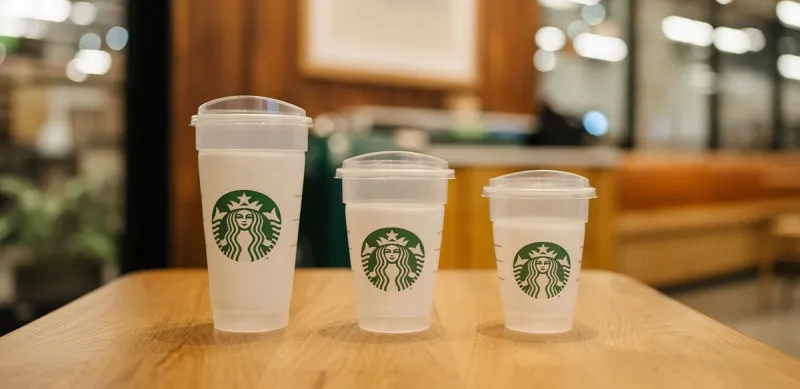 Tres vasos blancos de Starbucks de distintos tamaños, con tapas transparentes y el logotipo verde, colocados sobre una mesa de madera dentro de una cafetería.
