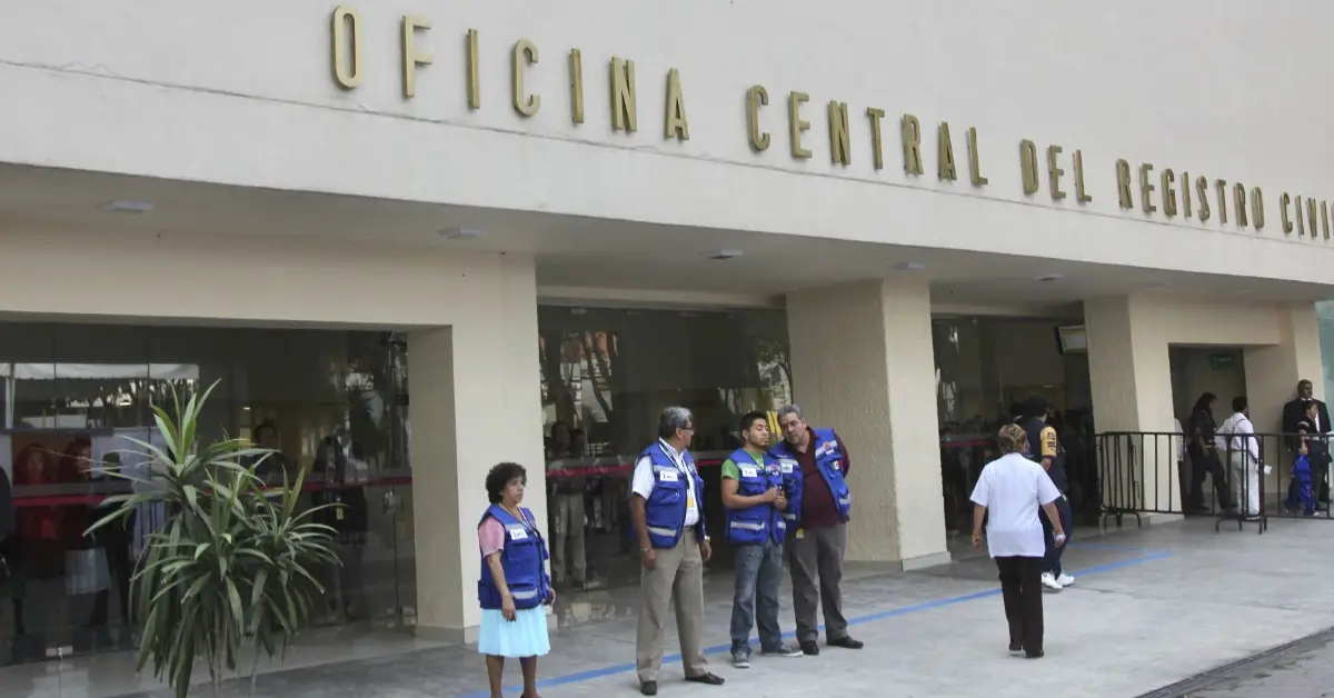 Fachada de edificio color beige con letras de "Oficina Central del registro Civil" y cuatro hombres y una mujer con chaleco azul .jpg