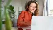 Young happy woman student using laptop watching webinar writing at home.