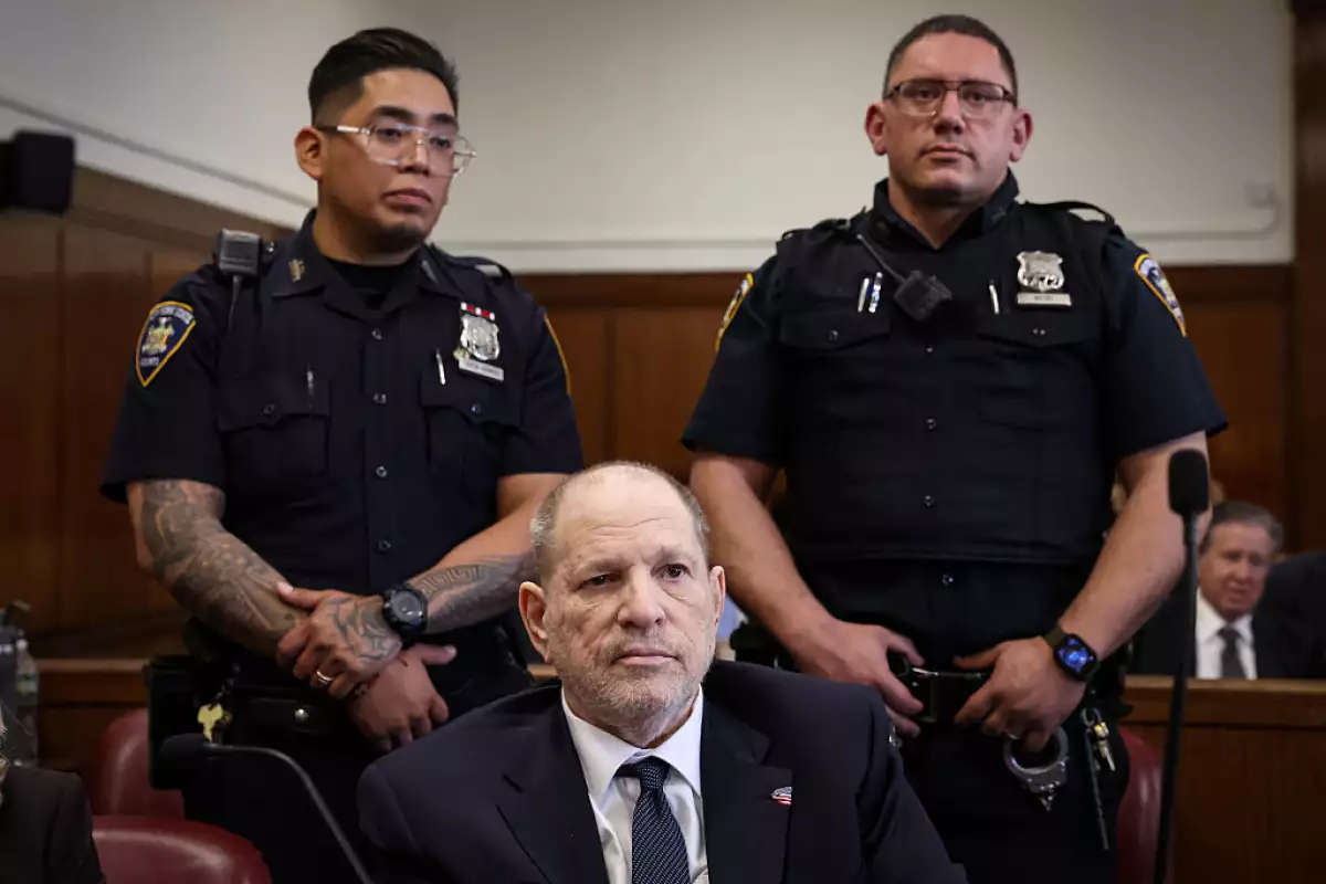 Foto de Harvey Weinstein vestido de traje y corbata con dos policías vigilándolo detrás durante su juicio.