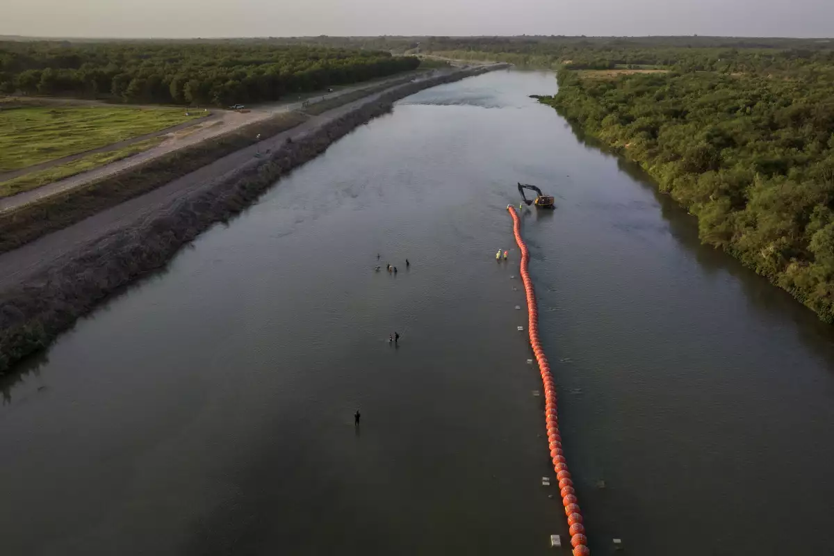 El huerto de nueces de Heavenly Farms se ve a la izquierda mientras las familias migrantes navegan por el río Río Grande pasando por trabajadores y boyas mientras buscan un punto de entrada a los Estados Unidos desde México en Eagle Pass, Texas, EE. UU., el 27 de julio de 2023.