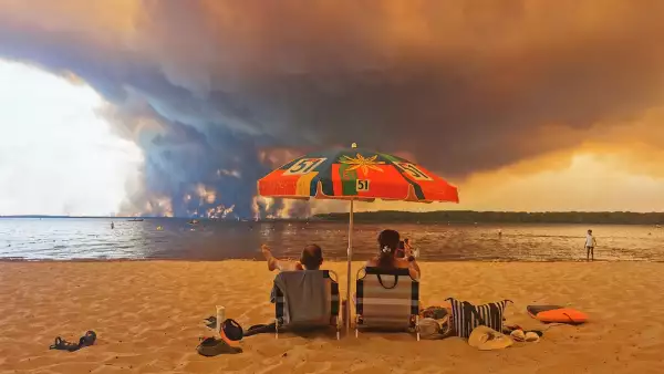 Los bañistas se sientan en la playa de Moutchic en Lacanau, al suroeste de Francia, el 24 de julio de 2026, mientras las nubes de humo se elevan al cielo a partir de un incendio forestal.