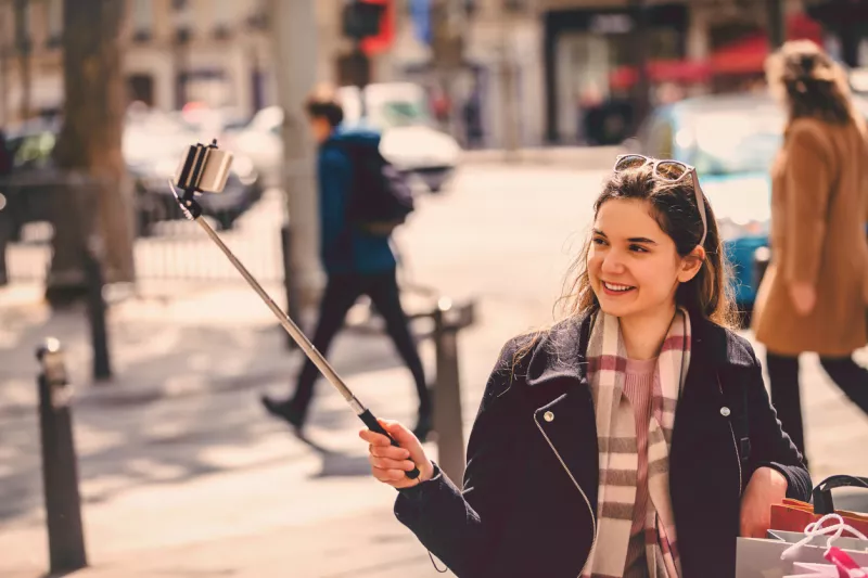 Vlogger joven mujer francesa tomando un video con un palo de selfie y un teléfono inteligente para publicar en las redes sociales un banco público sentado en la calle de París, Francia