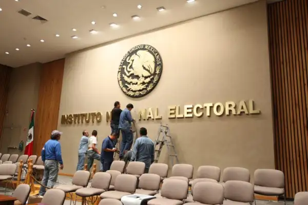 Bancas de color café en un auditorio, siete trabajadores, dos en escaleras, letras del Instituto Nacional Electoral y una bandera mexicana.jpg