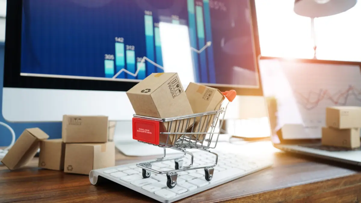 Cajas de papel en carrito de compras  sobre un teclado de computadora, al fondo un gráfico de crecimiento en la pantalla de la computadora.