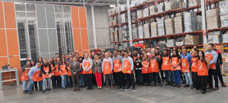 Personal de Home Depot vistiendo los mandiles de dependientes de la cadena en color naranja con el logo de la cadena en color blanco. Están en uno de los centros de distribución de la empresa por lo que hay algunos racks en la parte del fondo. 