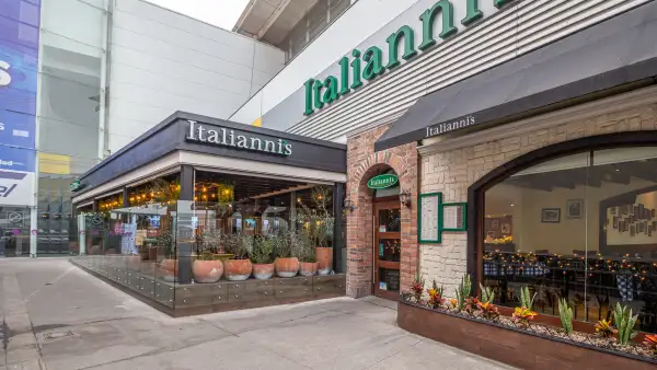 :Fachada de un restaurante Italianni’s en un centro comercial, con ventanales amplios, jardineras, detalles de ladrillo y piedra, y letreros con el nombre de la marca.