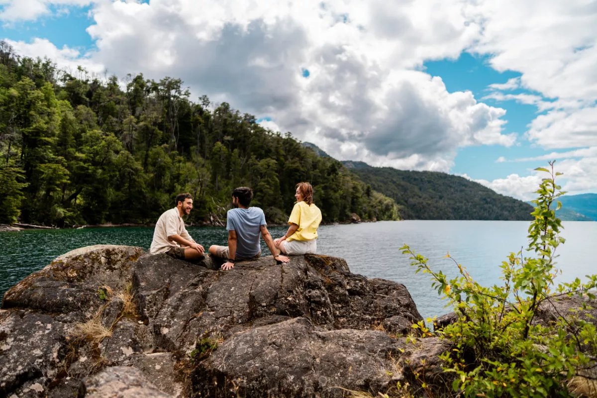 Foto de tres personas conversando sentadas sobre una roca frente a un lago de Patagonia, Argentina.