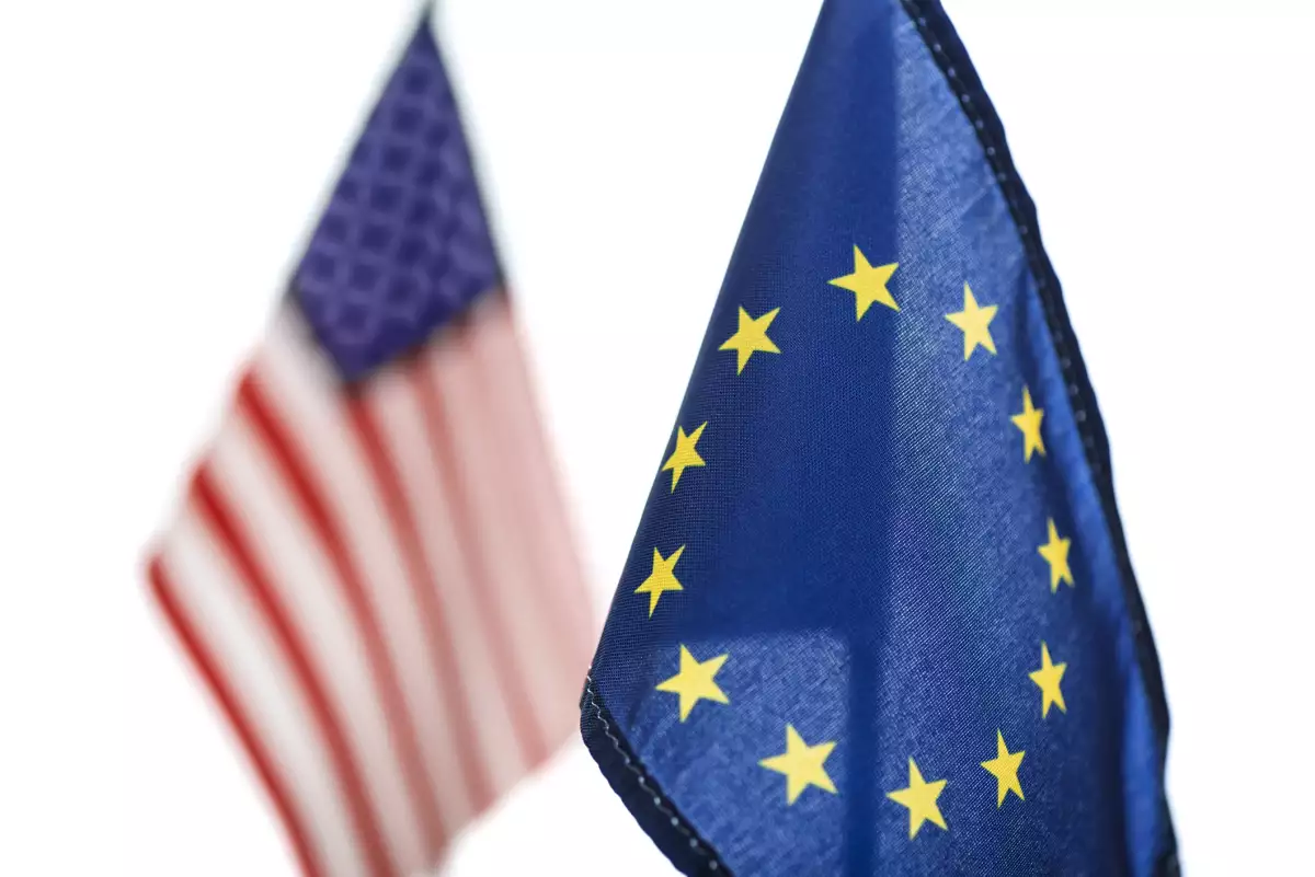 Primer plano de la bandera de la Unión Europea con la bandera estadounidense fuera de foco sobre un fondo blanco. Concepto de negociaciones y diplomacia. Imagen principal del artículo 'La Unión Europea se alista para responder a las amenazas comerciales de Trump, pero es la más vulnerable'.