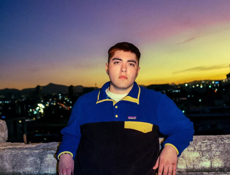 Foto de Kevin Kaarl recargado en una barda, lleva playera de manga larga y se ve el atardecer detrás.