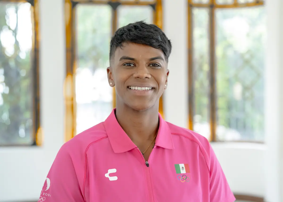 Foto medium shot de Prisca Awiti sonriendo, viste playera rosa mexicano con la bandera de México bordada, lleva cabello corto y aretes pequeños y discretos.