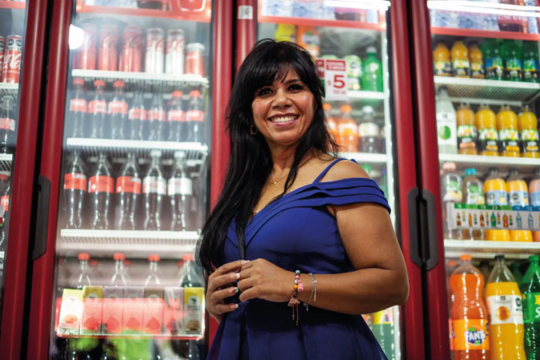 En primer plano aparece una tendera mexicana y en la parte de atrás un refrigerador lleno de productos Coca-Cola.