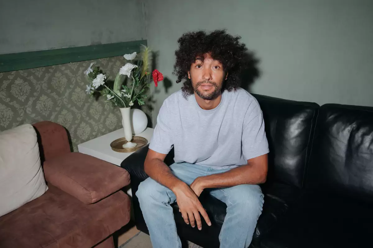 Foto del DJ &Me, sentado en una sala de piel negra, en una habitación de paredes verdes, él viste playera gris y jeans.