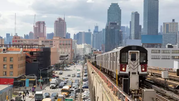 Tren acercándose a la estación de metro elevada en Queens, Nueva York. Los edificios financieros y el horizonte de Nueva York se ven en el fondo, a la izquierda abajo se puede ver una calle concurrida llena de coches en hora punta, cielo nublado, dramático, EE. UU.