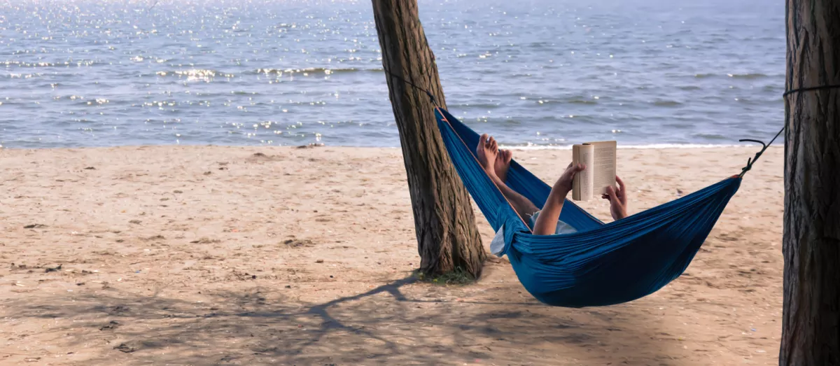 Foto de un hombre en una hamaca leyendo un libro en la playa frente al mar