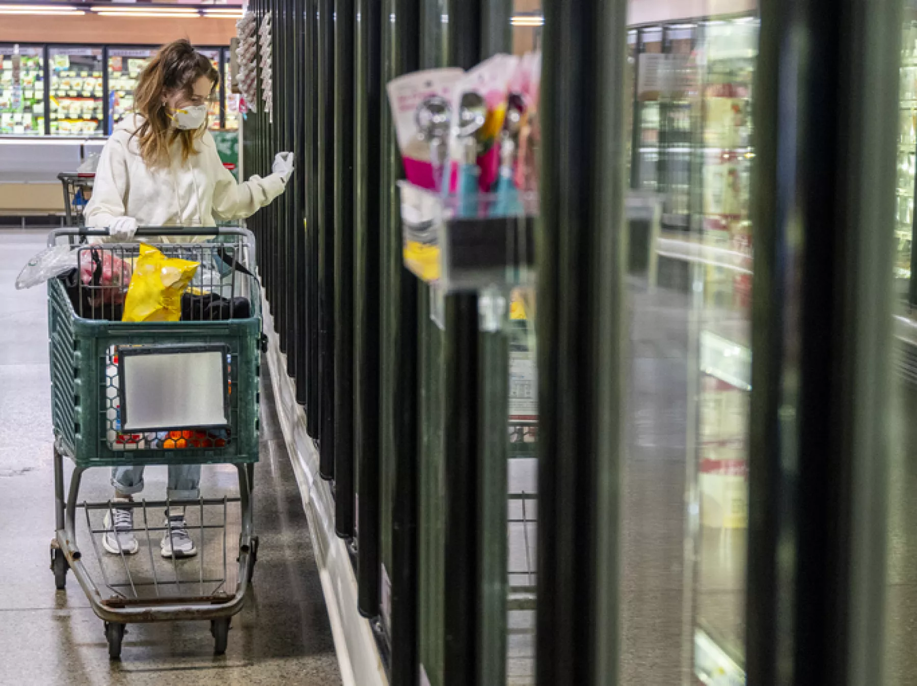 A young woman wearing a protective mask and gloves shopping in a time of virus pandemic, buying food supplies - processed frozen foods.