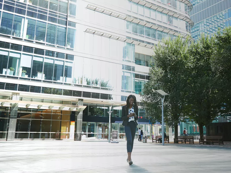 Imagen de una joven que revisa su celular mientras camina afuera de un gran edificio.