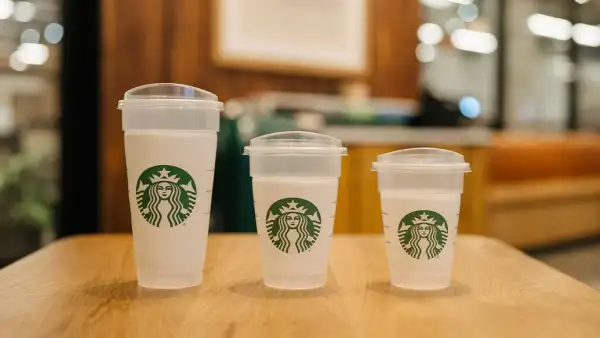 Tres vasos blancos de Starbucks de distintos tamaños, con tapas transparentes y el logotipo verde, colocados sobre una mesa de madera dentro de una cafetería.