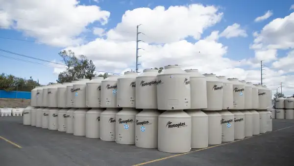 Un espacio exterior de almacenamiento donde se observan decenas de grandes tanques cilíndricos blancos de almacenamiento de agua, colocados en varias hileras y niveles. Los depósitos muestran el logotipo de Rotoplas y una capacidad visible de 1,100 litros. Los tanques ocupan la mayor parte de la imagen y están sobre un amplio pavimento oscuro con líneas amarillas, mientras que al fondo se distinguen árboles, una barda azul, postes y cables eléctricos. El cielo azul intenso, cubierto parcialmente por grandes nubes blancas, contrasta con los depósitos y el entorno industrial.