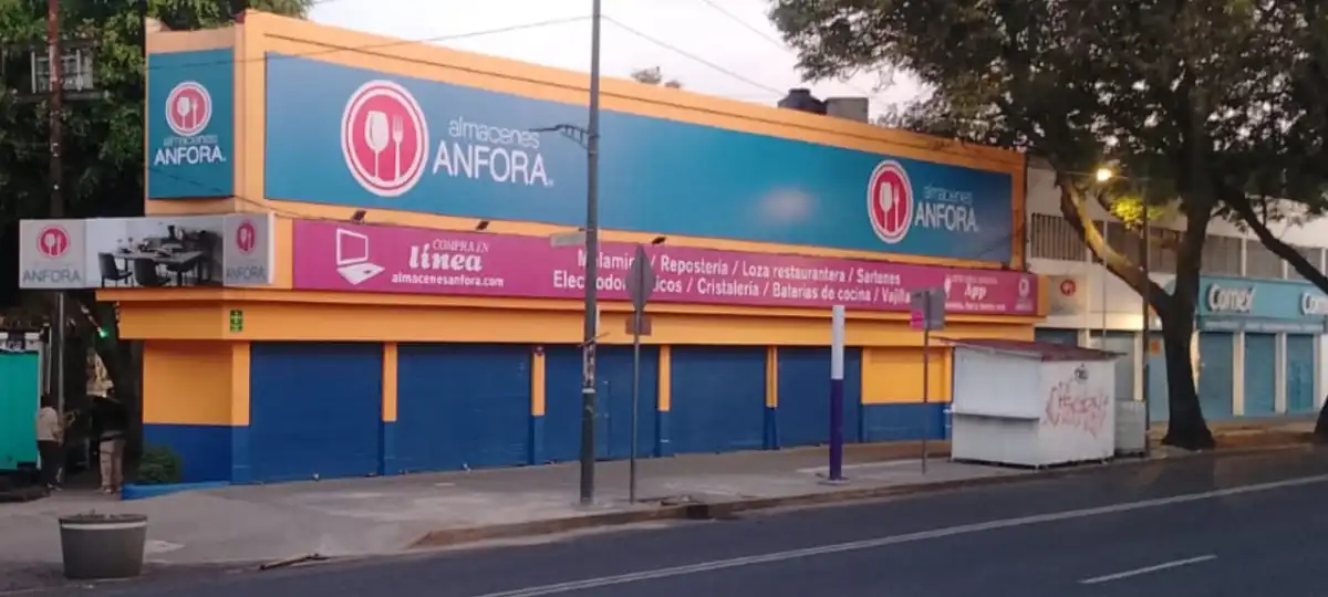 Sucursal de Almacenes Ánfora sobre una avenida, con una fachada de dos niveles en azul y naranja, el logotipo de la empresa visible en distintos puntos y anuncios de categorías como repostería, loza restaurantera, sartenes, electrodomésticos, cristalería, baterías de cocina y vajillas. La entrada tiene cortinas metálicas cerradas y al frente se observan una banqueta, postes, árboles y un pequeño puesto, en una escena urbana.
