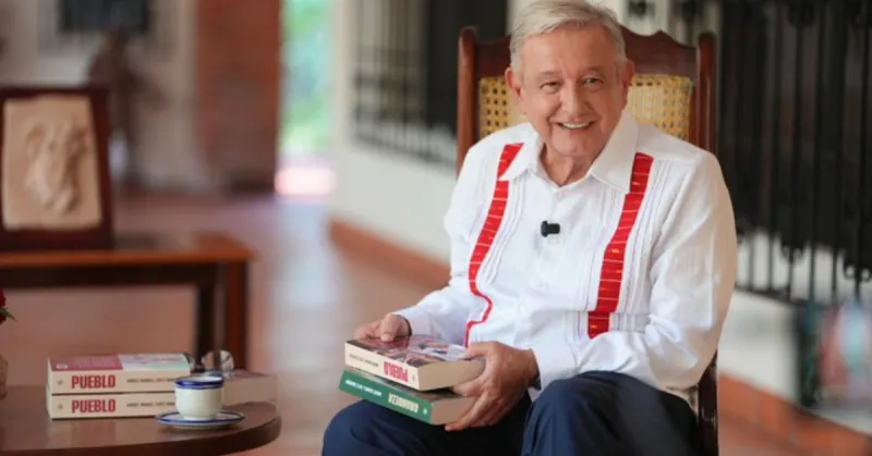 Andrés Manuel López Obrador sostiene dos libros en sus manos, viste guayabera blanca con vivos rojos y pantalón azul.jpg