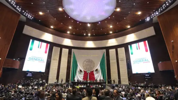 Legisladores al interior de la Cámara de Diputados, donde al fondo se observan dos banderas de México.