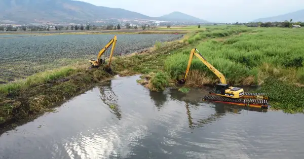 Un cuerpo de agua en medio de un área verde y dos grúas de color amarillo, y rodeado de montañas y cielo azul.jpg
