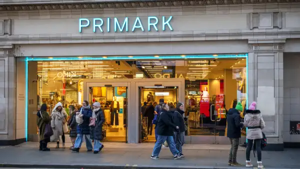 Fachada de una tienda Primark con un letrero de letras azul turquesa iluminadas sobre una entrada amplia. A través de los ventanales se observa el interior de la tienda, con luces cálidas amarillas, exhibidores de ropa y maniquíes. Varias personas con abrigos oscuros y de colores claros caminan o se detienen frente al establecimiento, ubicado en un edificio de piedra gris.