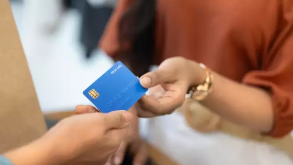 El brazo de una mujer sosteniendo una tarjeta de crédito para pagar una compra, junto con la mano de otra persona que acepta el pago en una terminal bancaria