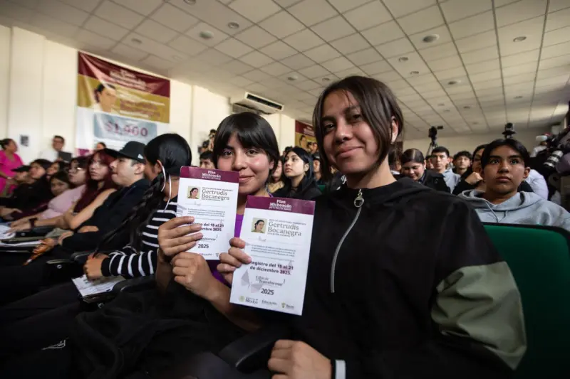 Dos estudiantes de preparatoria sentadas y rodeadas de más estudiantes muestran la tarjeta en la que reciben el pago de la beca Benito Juárez.