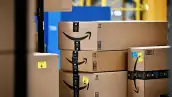 Paquetes de cartón de Amazon, algunos con cinta de embalaje de Prime y etiquetas de envío, apilados sobre una banda transportadora dentro de un centro logístico, con estructuras y maquinaria industrial al fondo.
