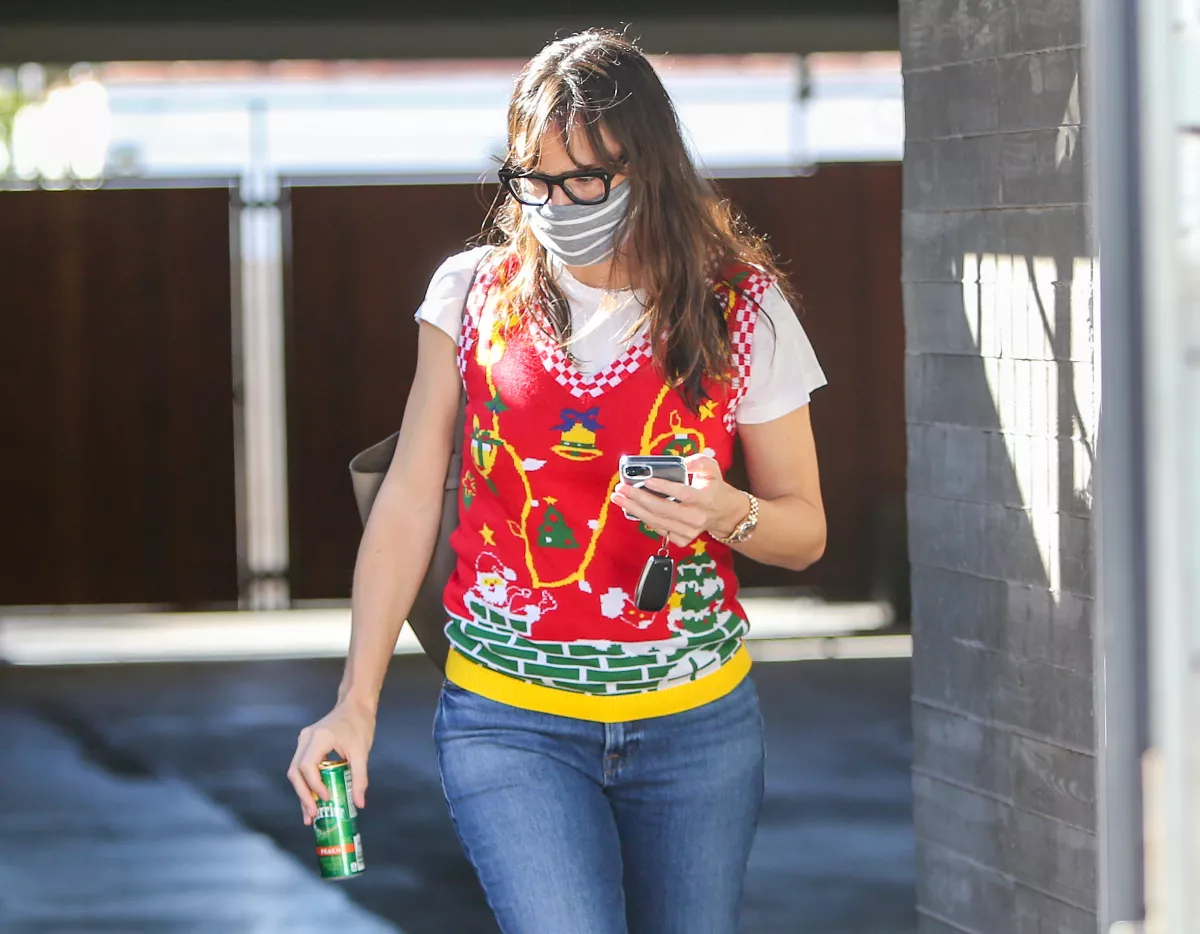 Foto de Jennifer Garner con cubrebocas, lentes de pasta, el celular en la mano y vistiendo una t-shirt blanca y encima un chaleco navideño y jeans.