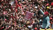 El presidente Luiz Inácio Lula da Silva saluda a una multitud de simpatizantes durante un acto de campaña en Brasil. Los asistentes, vestidos principalmente de rojo, levantan sus teléfonos para fotografiarlo y portan banderas y carteles con el número 13, asociado a su candidatura. Lula viste una camisa azul claro y una gorra roja mientras avanza entre la concentración.