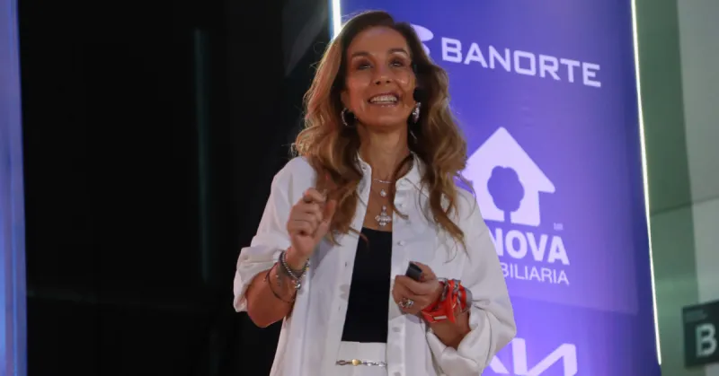 Carla Juan Chelala viste traje blanco y blusa negra. Sonríe, porta un micrófono alámbrico, detrás hay un cártel de Banorte en morado .jpg