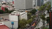 Edificios en lado izquierdo y derecho, al frente hay árboles y gente avanzando sobre una avenida, portan mantas.jpg